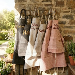 May include: A collection of personalized aprons hanging on a stone wall. The aprons are in various colors, including black, gray, and red, with vertical stripes. Each apron has a name embroidered on it in a contrasting color.