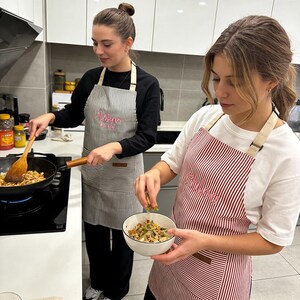 May include: Two people wearing striped aprons in a kitchen. One apron is gray and white striped, the other is red and white striped. Both aprons have the name "Chloe" embroidered on them in pink script. They are cooking.