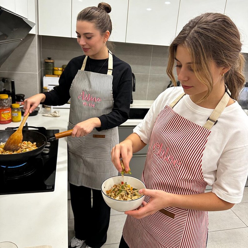 May include: Two people wearing striped aprons in a kitchen. One apron is gray and white striped, the other is red and white striped. Both aprons have the name "Chloe" embroidered on them in pink script. They are cooking.