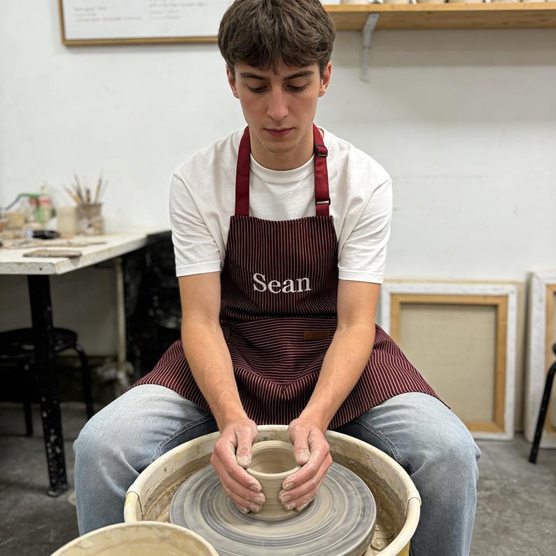 May include: A person working on a pottery wheel, shaping a clay pot. They are wearing a white t-shirt, blue jeans, and a burgundy striped apron with the name "Sean" embroidered on it. The pottery wheel is centered in the image.