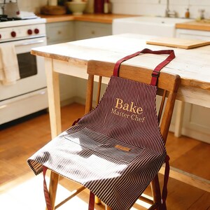 May include: A burgundy and white striped apron with the words "Bake Master Chef" in gold lettering. The apron is hanging on a wooden chair in a kitchen setting. The apron has a pocket and a tag.