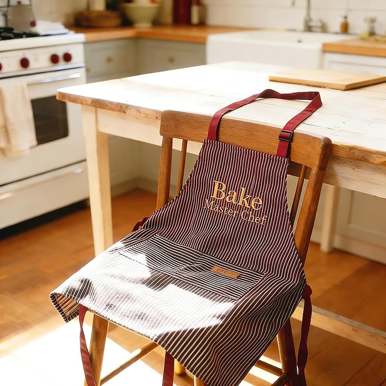 May include: A burgundy and white striped apron with the words "Bake Master Chef" in gold lettering. The apron is hanging on a wooden chair in a kitchen setting. The apron has a pocket and a tag.