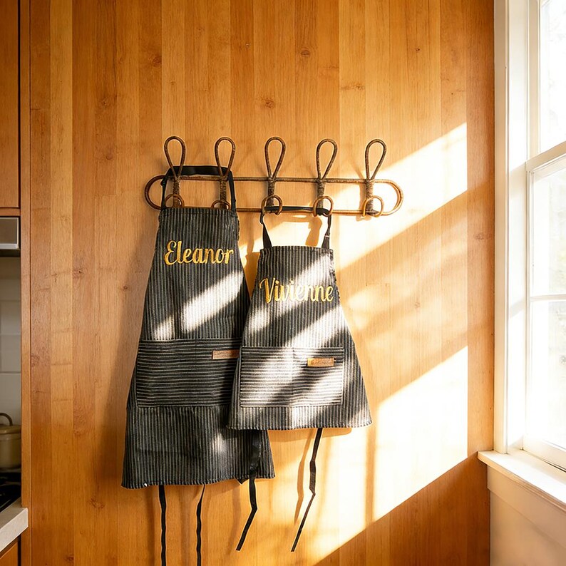 May include: Two black and gray striped aprons with the names "Eleanor" and "Vivienne" embroidered in gold thread. The aprons hang on a decorative metal rack against a wooden wall. The aprons have pockets and leather tags.