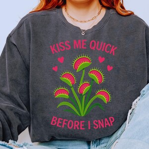 May include: Dark gray sweatshirt with a graphic of a Venus flytrap plant in pink and green, with the text "KISS ME QUICK BEFORE I SNAP" in pink. The design includes pink hearts.