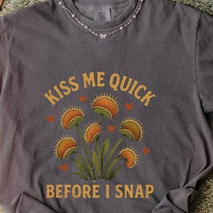 May include: Dark gray t-shirt featuring a graphic of Venus flytraps with the text "KISS ME QUICK BEFORE I SNAP" in gold. The design includes orange hearts and detailed illustrations of the plants. A casual, botanical-themed tee.