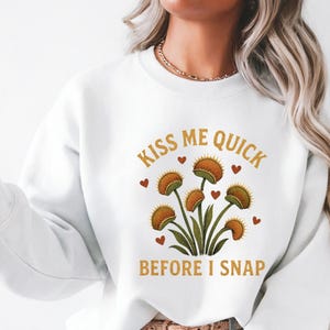 May include: White sweatshirt with a graphic of Venus flytraps and the text "Kiss Me Quick Before I Snap" in a playful font. The design features orange and green plants with red heart accents.