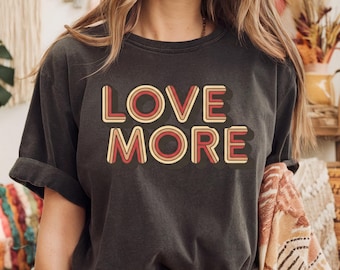 May include: A charcoal gray short-sleeve t-shirt featuring the text LOVE MORE printed in a layered, retro-inspired font. The letters are colored in shades of red, orange, and yellow with a dark drop shadow, creating a bold, multi-dimensional effect.