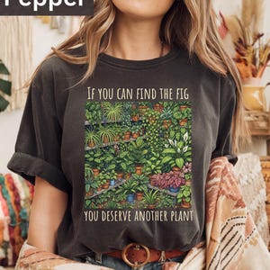 May include: Dark grey t-shirt featuring a botanical illustration of potted plants. The text on the shirt reads, "IF YOU CAN FIND THE FIG YOU DESERVE ANOTHER PLANT."