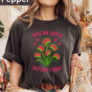 Puede incluir: Camiseta gris oscuro con el texto "KISS ME QUICK BEFORE I SNAP" en rosa. La camiseta presenta un gráfico de una Venus atrapamoscas con detalles rosas y verdes. La camiseta tiene mangas cortas y cuello redondo.