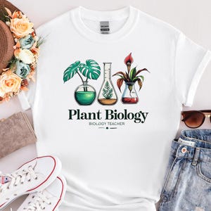 May include: A white t-shirt featuring a graphic of three laboratory glass vessels containing various plants. The text "Plant Biology" and "Biology Teacher" is printed in black below the illustrations. The shirt has a crew neck and short sleeves.