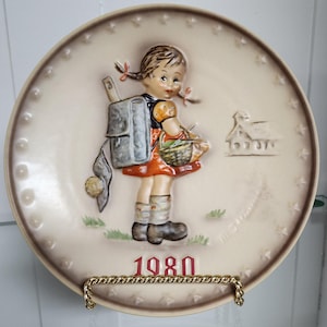 May include: A decorative ceramic plate featuring a young girl with pigtails, a backpack, and a basket. The girl is wearing a red dress and boots. The plate has a cream-colored background with a brown border and the year "1980" in red.