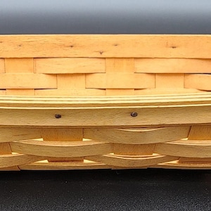 May include: A rectangular, light-colored woven basket with rounded corners. The basket is made of interwoven strips of wood, creating a textured surface. Small metal rivets secure the top rim. The basket is empty and sits on a dark surface.