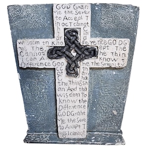 May include: A gray and white cross-shaped religious plaque with the Serenity Prayer inscribed. The cross features a Celtic knot design at the center and is set against a textured, gray background. The text is in a handwritten style.