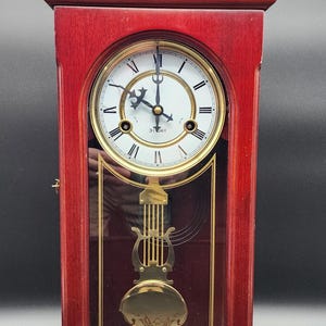May include: A wooden wall clock with a reddish-brown finish, featuring a white circular face with black Roman numerals and the text 31 DAY. A gold-colored lyre-shaped pendulum hangs behind a glass panel with gold decorative line accents.