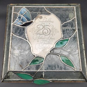 May include: A square stained glass piece with a silver frame. The design features a butterfly, green leaves, and a central panel with the text "Congratulations on 25 Silver Years Together."