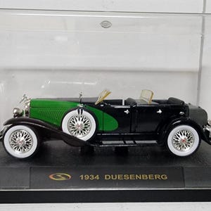 May include: A 1934 Duesenberg model car in a display case. The classic car is black with green accents and white-walled tires. The model sits on a black base with gold lettering.