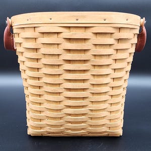 May include: A woven basket with a light brown color. The basket has a rectangular shape with a slightly curved top. It features brown leather handles attached to the sides. The basket is made of woven wood strips.