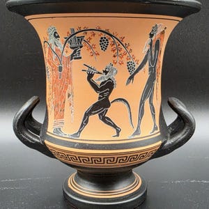 May include: A black and terracotta-colored ceramic vase featuring a wide rim, two curved handles, and a pedestal base. The exterior displays a black-figure style illustration of a bearded figure, a satyr playing a flute, and a grape vine.