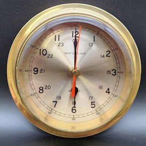 May include: A round ship's clock with a polished brass frame and a metallic gold face. The dial features black Arabic numerals for both 12-hour and 24-hour time formats, with the text SHIP'S CLOCK printed in black. It has black hour and minute hands and a thin red second hand.