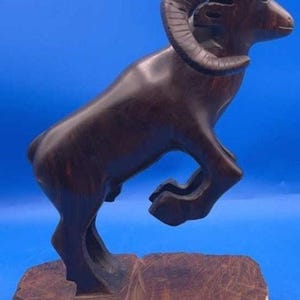 May include: A dark brown wooden sculpture of a ram in a leaping pose. The ram has large, curved horns and is mounted on a rough-hewn wooden base. The sculpture is set against a blue background.