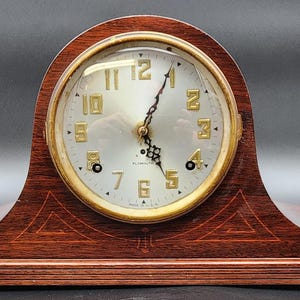 May include: A vintage wooden mantel clock with a light-colored face and gold-colored numerals. The clock has a gold-colored bezel and a dark brown wooden case. The clock face reads "HAMILTON". The clock is set against a dark background.