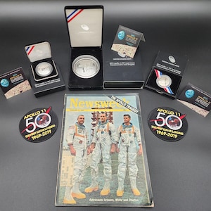 May include: An assortment of commemorative items related to the Apollo 11 mission. Includes coins in display boxes, patches with "Apollo 11 50" text, and a vintage "Newsweek" magazine cover featuring astronauts. The items are on a dark surface.