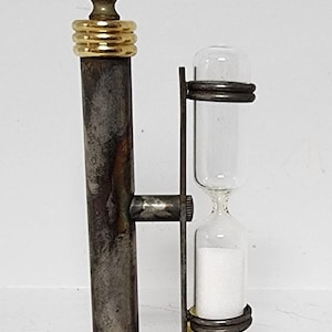 May include: A decorative hourglass featuring a clear glass bulb filled with white sand, mounted on a weathered metal post with gold-toned rings and a faceted crystal finial. The base is a rectangular metal block with a blue logo print.