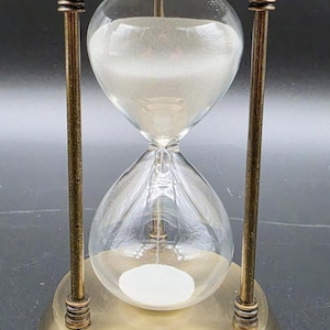 May include: An hourglass featuring a clear glass bulb filled with white sand, supported by a metallic frame with a brushed brass finish. The structure consists of a circular base and a matching top cap connected by three vertical pillars.