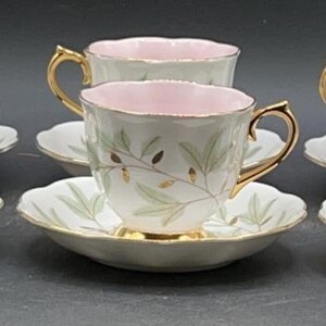 May include: A set of six vintage teacups and saucers. The cups have a pink interior, gold handles, and a white exterior with a green leaf design. The saucers are white with a scalloped edge and gold trim.