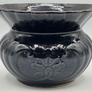 May include: A black ceramic vase with a glossy finish. The vase has a wide, fluted rim and a rounded body with a floral design. The vase is approximately 6 inches tall and 7 inches wide.