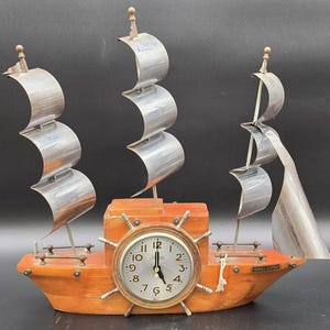 May include: A ship-shaped wooden clock featuring three silver-toned metal masts with curved sails. The clock face is centered within a ship's wheel design on the hull, displaying the text "SETH THOMAS" and "YANKEE CLIPPER".