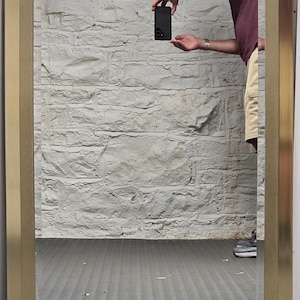 May include: A rectangular wall mirror featuring a wide, brushed gold-tone metal frame with mitered corners. The reflective glass surface shows a clear view of the surroundings, including a textured stone wall and a dark gray floor surface.