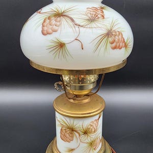 May include: Vintage oil lamp with a white glass shade and base, adorned with hand-painted pine cone and branch designs in brown and green. The lamp features brass accents and a clear glass chimney.