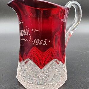 May include: A ruby red glass pitcher with a clear handle and base. The pitcher has etched details and the text "mit ~ 1903." The base features a detailed, cut-glass design.