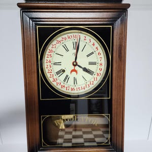 May include: A rectangular wall clock with a dark brown wooden frame. The clock face features black Roman numerals, a red crescent moon indicator, and a red outer ring numbered 1 to 31. The lower glass panel displays the word REGULATOR in gold.