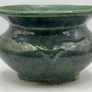 May include: A dark green ceramic bowl with a wide, flared rim and a rounded body. The bowl has a glossy, iridescent finish with subtle variations in color. The exterior features embossed floral or leaf-like patterns. The bowl sits on a light-colored base.