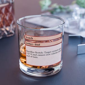 May include: A clear, lowball glass filled with amber liquid, possibly whiskey or scotch. A label on the glass reads "Scotch" and "Artifact - Food", with game instructions. The glass sits on a dark surface.