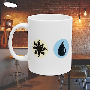 May include: White ceramic mug featuring two circular designs: a black sunburst on a cream background and a black teardrop on a light blue background. The mug has a large, curved handle and is set against a brick wall background.