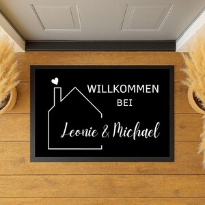 Könnte beinhalten: Schwarze rechteckige Fußmatte mit weißem Text und Hausumriss. Der Text lautet "WILLKOMMEN BEI Leonie & Michael" mit einem Herz über dem Haus. Die Matte liegt auf einem Holzboden.