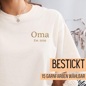 May include: Cream-colored t-shirt with the word "Oma" embroidered in gold thread, along with "Est. 2026". The shirt also features the text "BESTICKT" and "15 GARNFARBEN W&Auml;HLBAR" in a rectangular box.