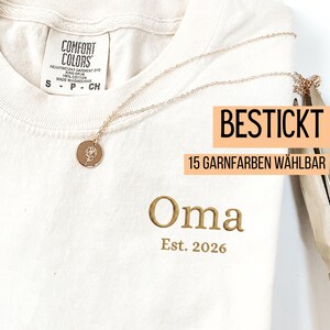 May include: Cream-colored t-shirt with the word "Oma" embroidered in gold, along with "Est. 2026". A gold necklace with a floral pendant rests on the shirt. The shirt tag reads "Comfort Colors". The text "BESTICKT 15 GARNFARBEN W&Auml;HLBAR" is also visible.