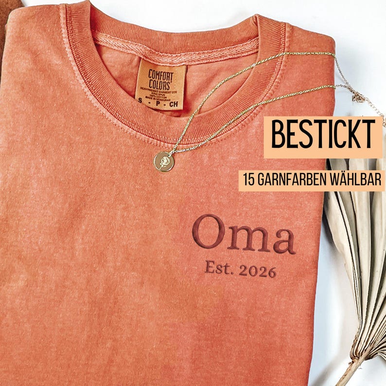 May include: A coral-colored Comfort Colors t-shirt with the embroidered word "Oma" and the year "Est. 2026". A gold necklace with a pendant is draped across the shirt. The text "BESTICKT" and "15 GARNFARBEN W&Auml;HLBAR" are also visible.