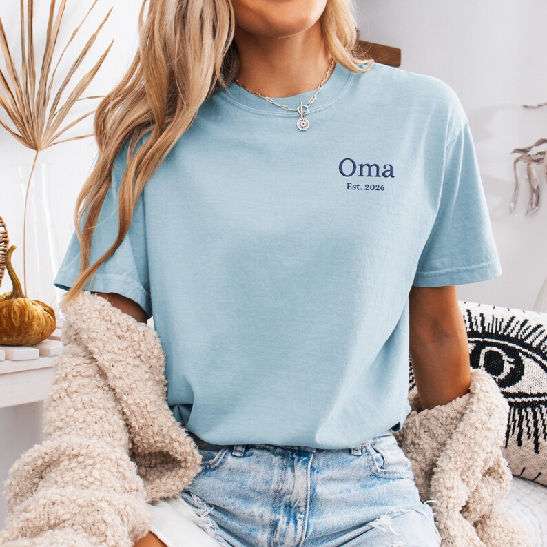 May include: Light blue t-shirt with the word "Oma" and the text "Est. 2026" printed in navy blue. The shirt is made of a soft material and has short sleeves. The shirt is worn with blue jeans.
