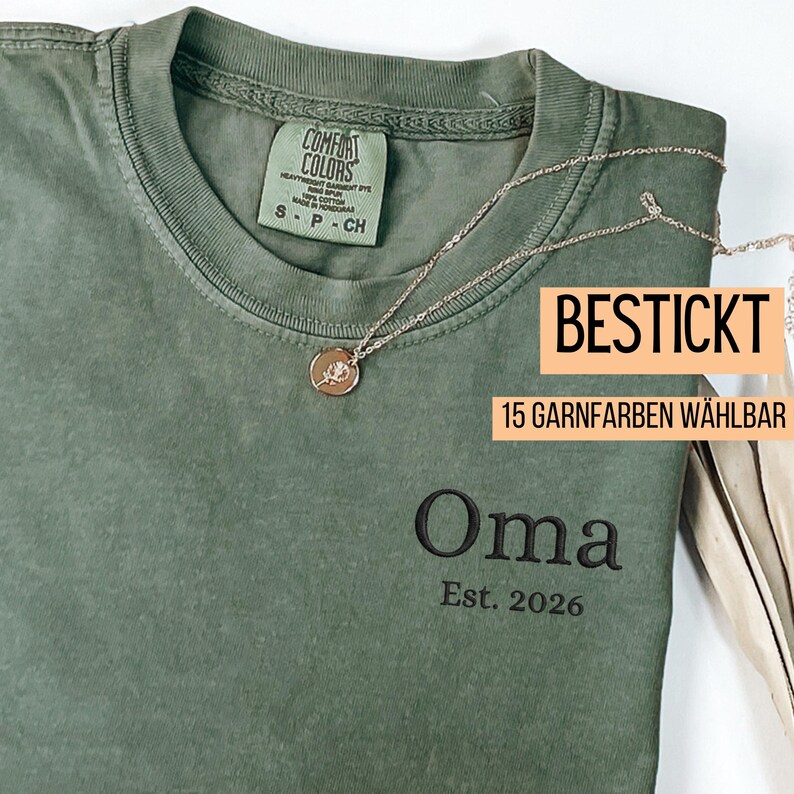 May include: Olive green t-shirt with the embroidered word "Oma" and "Est. 2026" in black thread. A silver necklace with a pendant is draped across the shirt. The shirt has a tag that says "COMFORT COLORS".