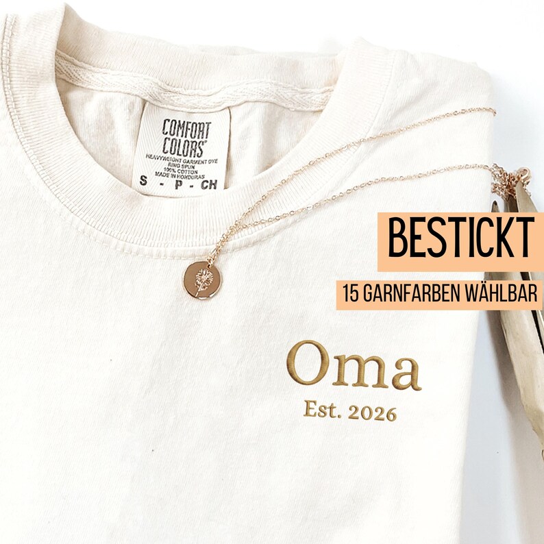 May include: Cream-colored t-shirt with the word "Oma" embroidered in gold, along with "Est. 2026". A gold necklace with a floral pendant rests on the shirt. The shirt tag reads "Comfort Colors". The text "BESTICKT 15 GARNFARBEN W&Auml;HLBAR" is also visible.