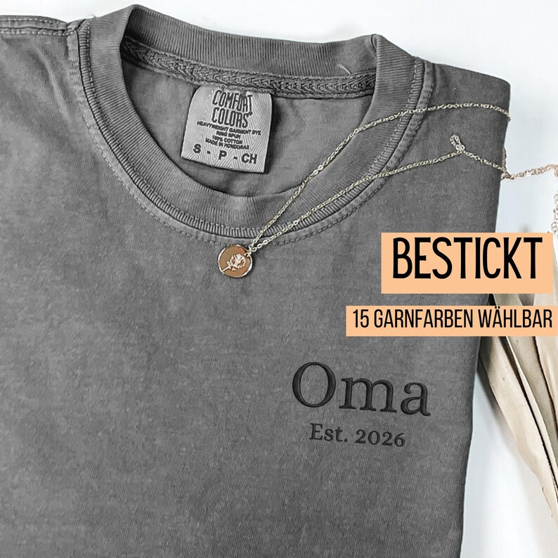May include: A gray Comfort Colors t-shirt with the embroidered word "Oma" and "Est. 2026". A silver necklace with a circular pendant is draped across the shirt. The text "BESTICKT" and "15 GARNFARBEN W&Auml;HLBAR" are also visible.
