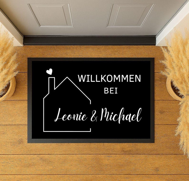 Könnte beinhalten: Schwarze rechteckige Fußmatte mit weißem Text und Hausumriss. Der Text lautet "WILLKOMMEN BEI Leonie & Michael" mit einem Herz über dem Haus. Die Matte liegt auf einem Holzboden.