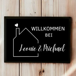 May include: Black rectangular sign with white text and a house outline. The sign reads "Willkommen bei Leonie & Michael" with a heart above the house outline. The sign is set against a wooden background.