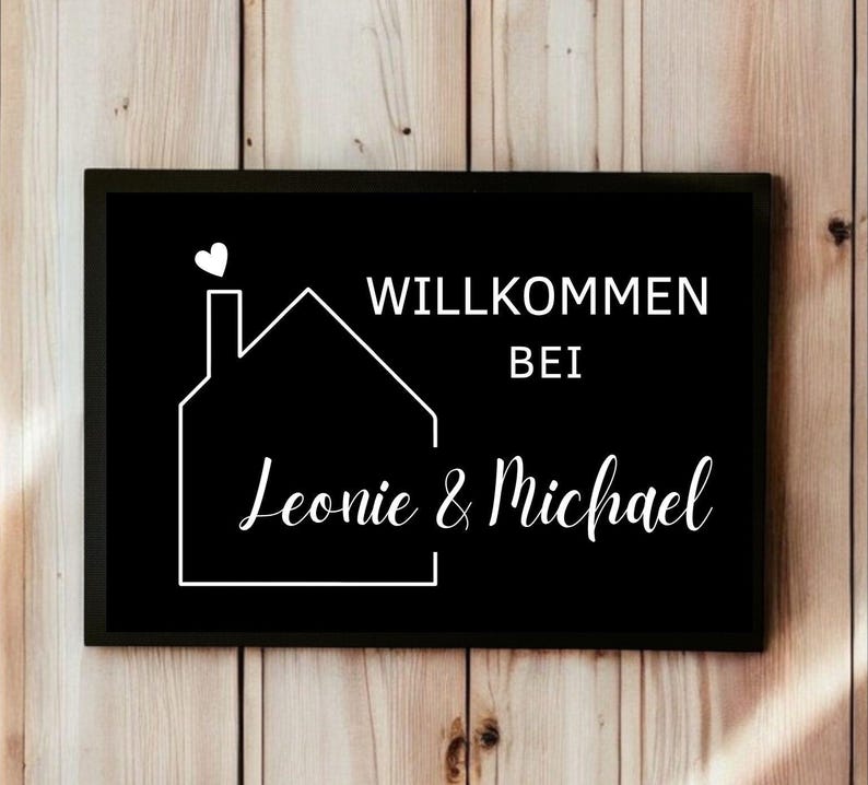May include: Black rectangular sign with white text and a house outline. The sign reads "Willkommen bei Leonie & Michael" with a heart above the house outline. The sign is set against a wooden background.