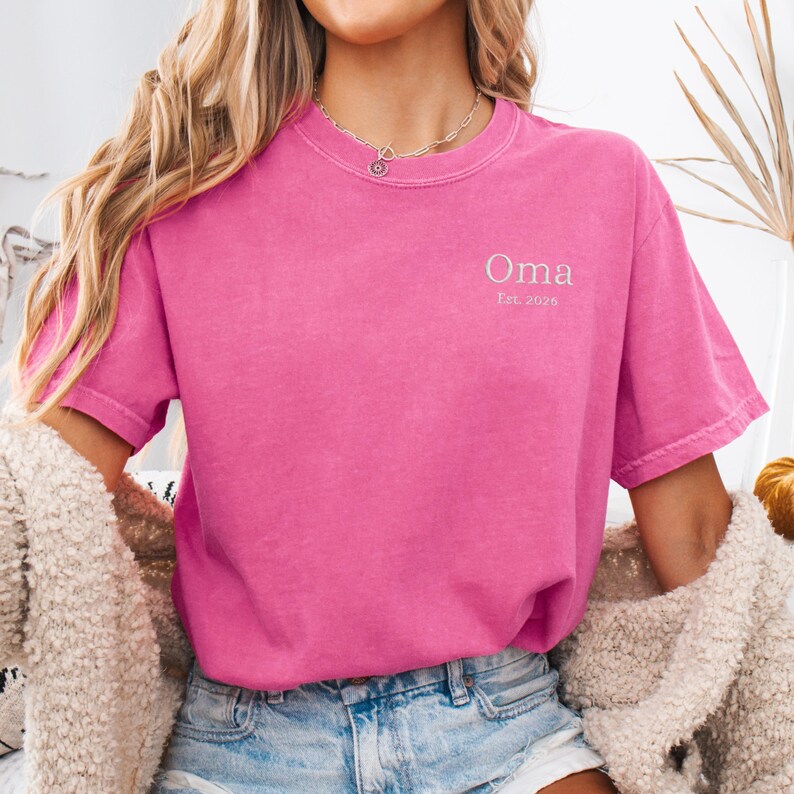 May include: A bright pink t-shirt with the word "Oma" embroidered in white, along with "Est. 2026". The shirt has a classic crew neck and short sleeves. The fabric appears soft and slightly faded.